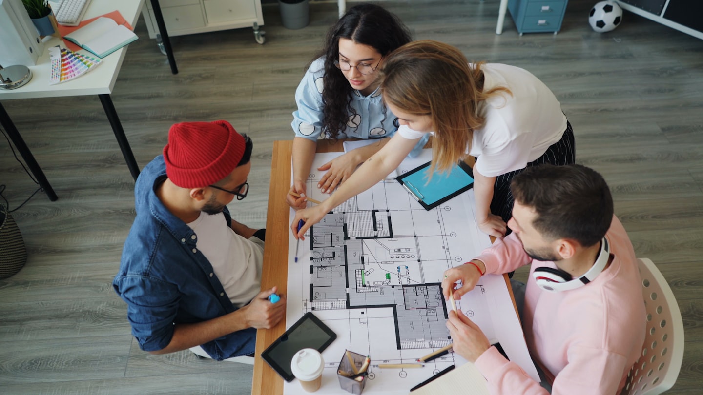Four designers collaborate over an architectural blueprint on a table.